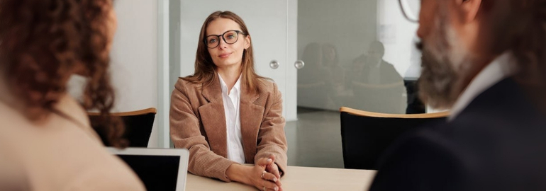 A woman wearing glasses engages in a job interview at a table with two men, responding to common interview questions.
