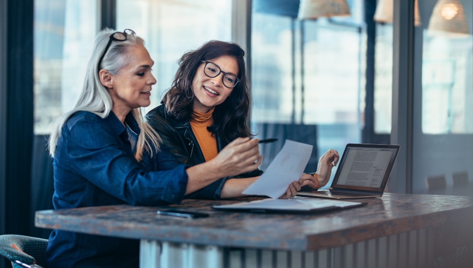 Two female professionals appearing to be having a work discussion with a laptop and paperwork at a desk