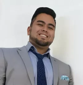 A man in an executive wear smiling while leaning on his side to a wall in a professional portrait shot.