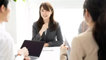 Four people in business attire are having a meeting at a bright office table. One woman is smiling and speaking while others listen, some holding laptops or tablets.