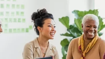 Four women are smiling and engaged in conversation in a bright office setting.