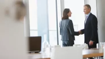 A man and woman shaking hands in a conference room, indicating a formal agreement or partnership.