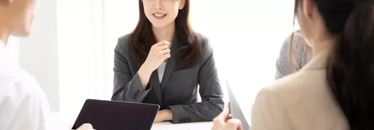 Four people in business attire are having a meeting at a bright office table. One woman is smiling and speaking while others listen, some holding laptops or tablets.